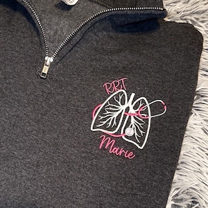 May include: Dark gray quarter-zip sweatshirt with an embroidered design. The design features a white lung illustration with pink accents and the text "RRT Marie". The sweatshirt has a silver zipper.