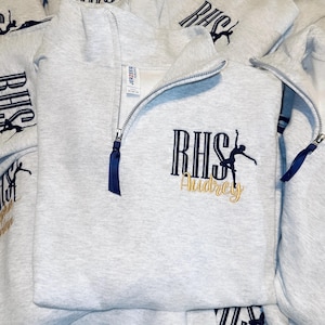May include: A light gray quarter-zip sweatshirt featuring a dark blue embroidered logo with the letters RHS and a silhouette of a dancer. The name Audrey is embroidered in gold script below the logo. The zipper pull is a dark blue ribbon.