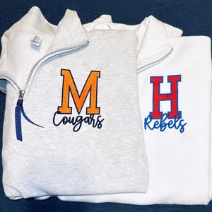 May include: Two quarter-zip sweatshirts. The left is light gray with an orange embroidered letter M and the word Cougars in navy blue. The right is white with a red embroidered letter H and the word Rebels in blue. Both have silver zippers.