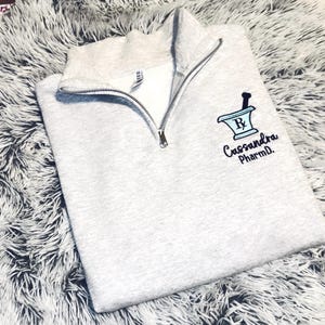 May include: A light gray quarter-zip sweatshirt with a silver zipper. The sweatshirt features a blue and black mortar and pestle design with the text "Cassandra PharmD." The garment is folded on a fluffy, light gray surface.