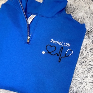 May include: A royal blue 1/4 zip sweatshirt with a black embroidered stethoscope, heartbeat, and heart design. The text "Rachel, LPN" is embroidered on the sweatshirt.