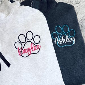 May include: Two quarter-zip sweatshirts, one light gray and one dark gray, each embroidered with a paw print design. The light gray sweatshirt has the name "Kaylee" embroidered in pink, while the dark gray one has "Ashley" in blue.