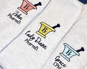 Könnte beinhalten: Nahaufnahme von drei hellgrauen Sweatshirts. Jedes Sweatshirt hat ein gesticktes Mörser- und Stößel-Design in verschiedenen Farben: Rot, Gelb und Blau. Die Namen "Zoha PharmD", "Lady Diane, PharmD" und "Grace CPhT" sind darunter gestickt.