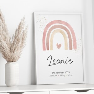 Peut inclure: Une impression d'arc-en-ciel aquarelle avec un sch&eacute;ma de couleurs rose, beige et marron. L'arc-en-ciel a un petit c&oelig;ur au centre. L'impression comprend le nom "Leonie", la date "09. Februar 2025", et l'heure "22:18Uhr", le poids "3315g", et la longueur "52cm".