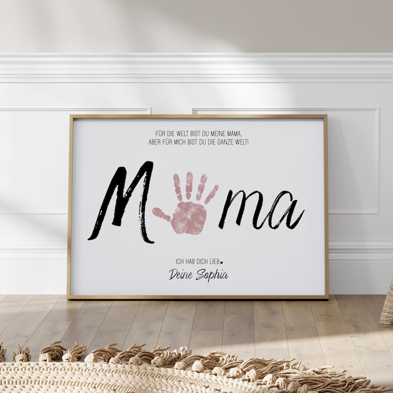 Puede incluir: Impresi&oacute;n enmarcada con la palabra "Mama" en escritura negra, con una huella de mano rosa. La impresi&oacute;n incluye texto en alem&aacute;n: "Para el mundo eres mi mam&aacute;, pero para m&iacute; eres todo el mundo!" y "Te amo, Tu Sophia."