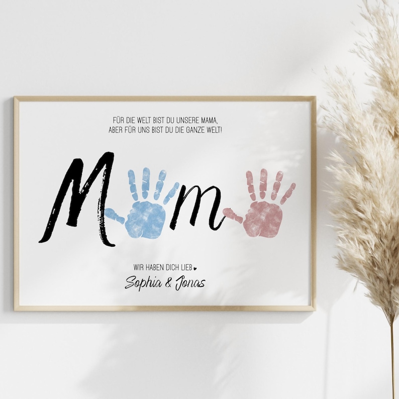 Puede incluir: Un cuadro enmarcado con la palabra "Mum" en escritura negra, con huellas de manos azules y rosas. La impresi&oacute;n incluye texto en alem&aacute;n, con los nombres "Sophia & Jonas" en la parte inferior. El marco es de color madera clara.