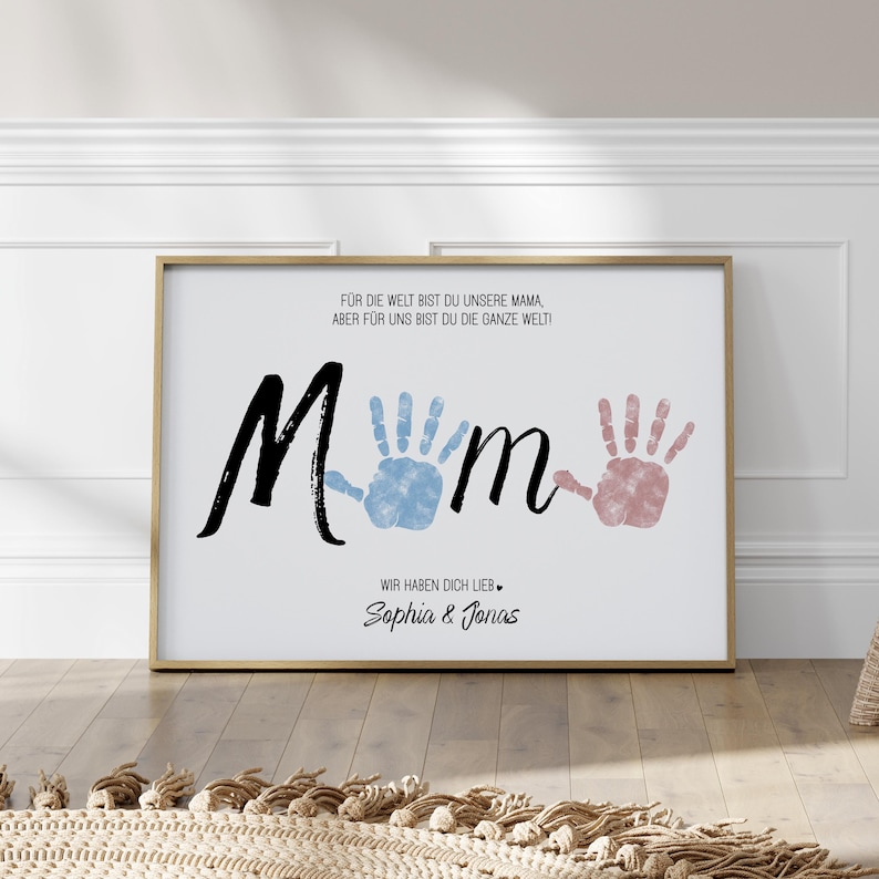 Puede incluir: Impresi&oacute;n enmarcada con la palabra "Mam&aacute;" en escritura negra, huellas de manos azules y rosas, y texto en alem&aacute;n. El marco dorado rodea un fondo blanco con el texto "&iexcl;Para el mundo eres nuestra mam&aacute;, pero para nosotros eres todo el mundo!" y nombres.