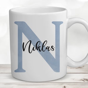 Könnte beinhalten: Weiße Keramiktasse mit einem blauen Aquarellbuchstaben N und dem Namen Niklas in schwarzer Kursivschrift.