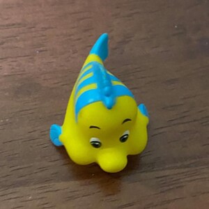 Pode incluir: Estatueta amarela e azul de Linguado do filme da Disney, A Pequena Sereia. A estatueta tem um corpo amarelo com listras e barbatanas azuis. O peixe tem olhos grandes e expressivos e uma express&atilde;o sorridente. A estatueta &eacute; um item de colecionador.