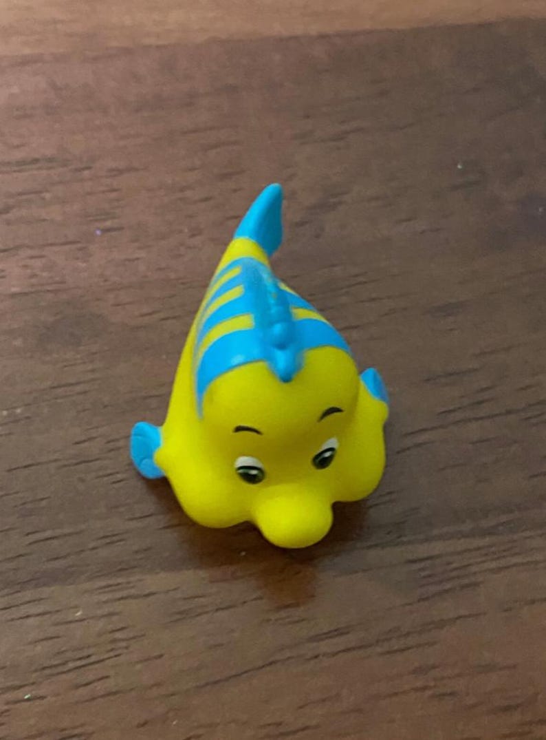 Pode incluir: Estatueta amarela e azul de Linguado do filme da Disney, A Pequena Sereia. A estatueta tem um corpo amarelo com listras e barbatanas azuis. O peixe tem olhos grandes e expressivos e uma express&atilde;o sorridente. A estatueta &eacute; um item de colecionador.