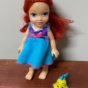 Pode incluir: Uma pequena boneca com cabelo ruivo, vestida com um vestido azul com detalhes roxos e uma gola branca. Um peixe de brinquedo amarelo com barbatanas azuis est&aacute; ao lado da boneca. A boneca tem olhos azuis e est&aacute; em p&eacute;.