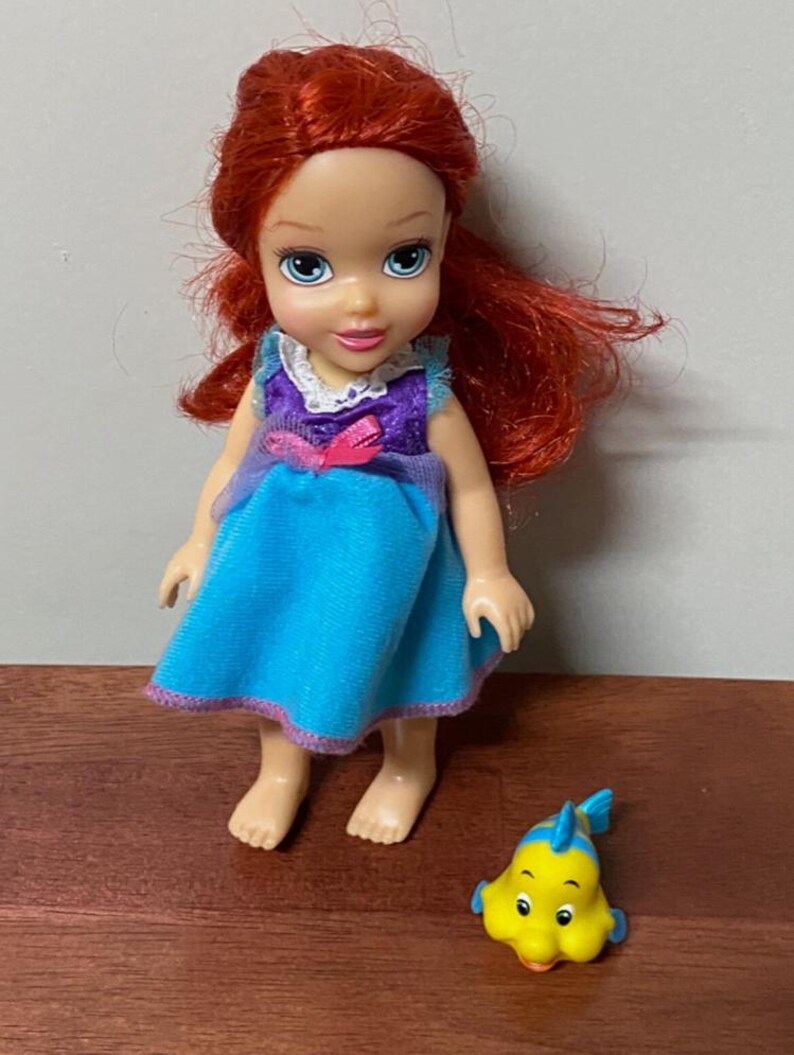 Pode incluir: Uma pequena boneca com cabelo ruivo, vestida com um vestido azul com detalhes roxos e uma gola branca. Um peixe de brinquedo amarelo com barbatanas azuis est&aacute; ao lado da boneca. A boneca tem olhos azuis e est&aacute; em p&eacute;.
