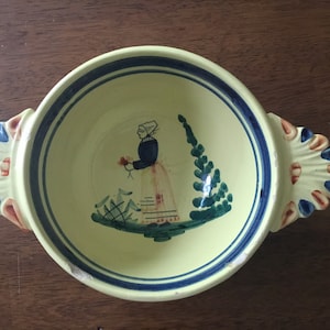 May include: A yellow ceramic bowl with blue and red painted details. The bowl has two handles and features a painted scene of a woman in a blue dress and white head covering holding a flower.