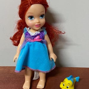 Pode incluir: Uma boneca de uma personagem com cabelo ruivo, vestida com um vestido azul com detalhes roxos. Uma figura de peixe amarelo e azul est&aacute; ao lado da boneca. A boneca est&aacute; sobre uma superf&iacute;cie castanha.