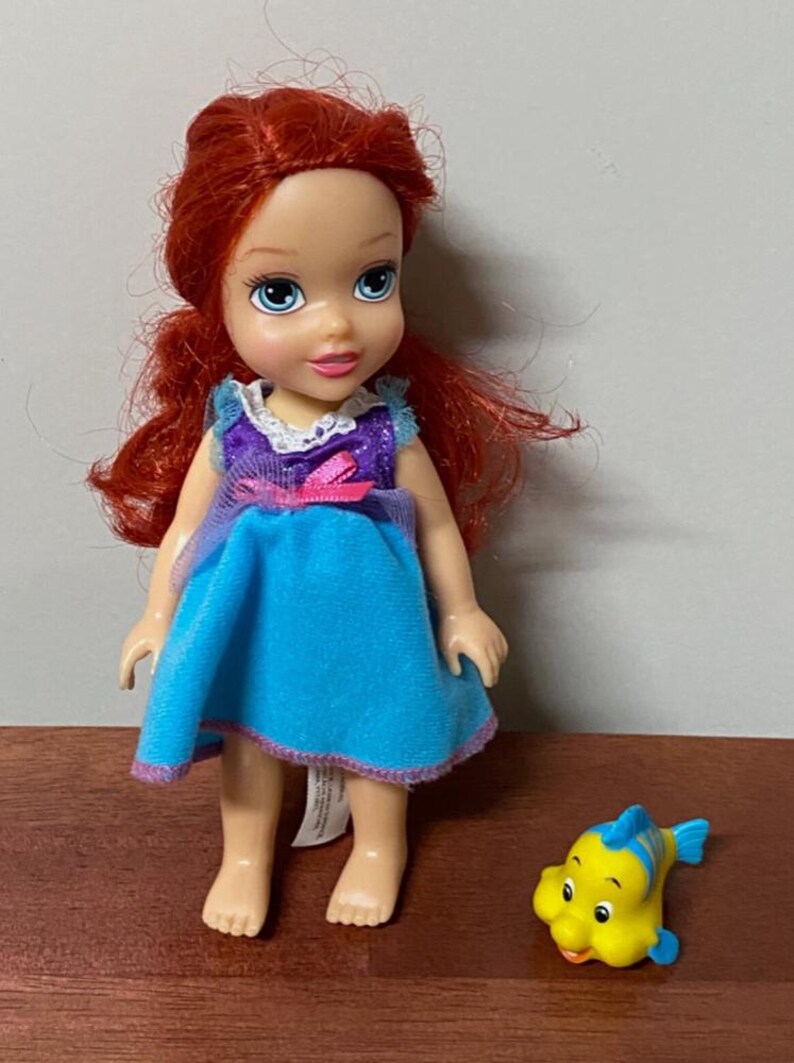 Pode incluir: Uma boneca de uma personagem com cabelo ruivo, vestida com um vestido azul com detalhes roxos. Uma figura de peixe amarelo e azul est&aacute; ao lado da boneca. A boneca est&aacute; sobre uma superf&iacute;cie castanha.