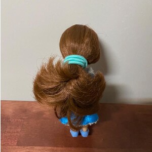 Pode incluir: Uma pequena boneca com cabelo castanho preso com um el&aacute;stico azul claro. A boneca est&aacute; vestida com um vestido azul e sapatos azuis. A boneca est&aacute; sobre uma superf&iacute;cie de madeira.