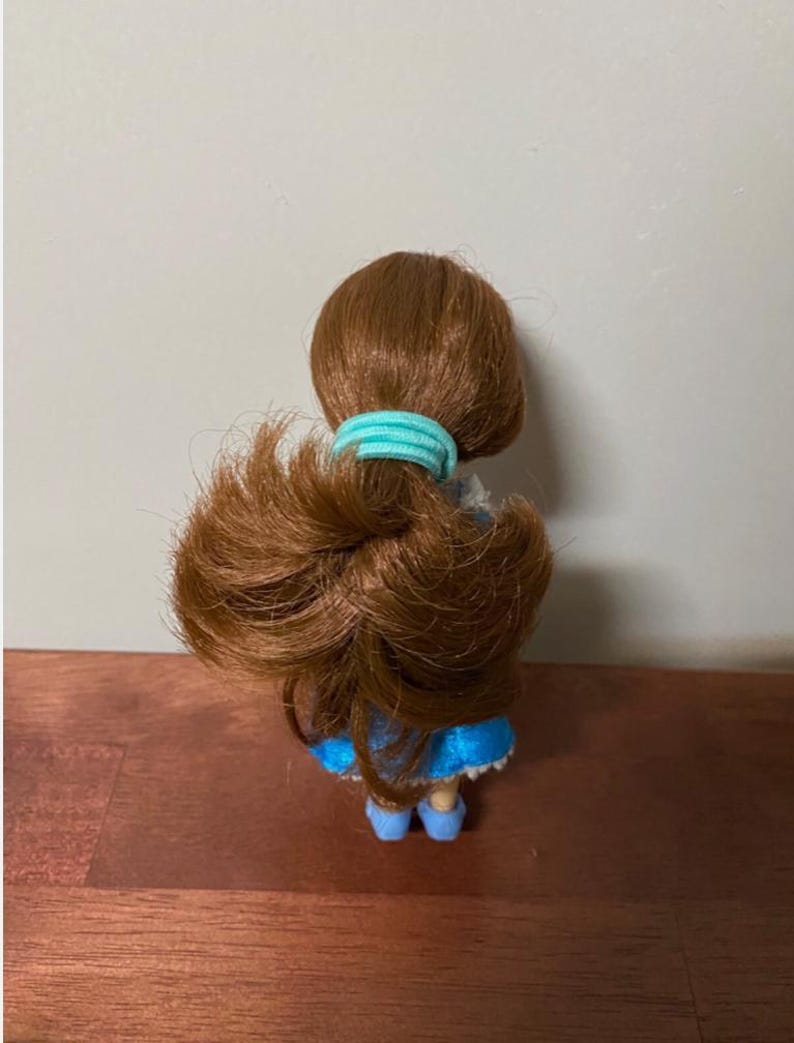 Pode incluir: Uma pequena boneca com cabelo castanho preso com um el&aacute;stico azul claro. A boneca est&aacute; vestida com um vestido azul e sapatos azuis. A boneca est&aacute; sobre uma superf&iacute;cie de madeira.