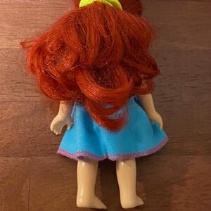 Pode incluir: Uma pequena boneca com cabelo vermelho vivo penteado em cachos soltos, preso com um el&aacute;stico verde n&eacute;on. A boneca usa um vestido azul claro com acabamento roxo. As pernas e os p&eacute;s da boneca s&atilde;o vis&iacute;veis.