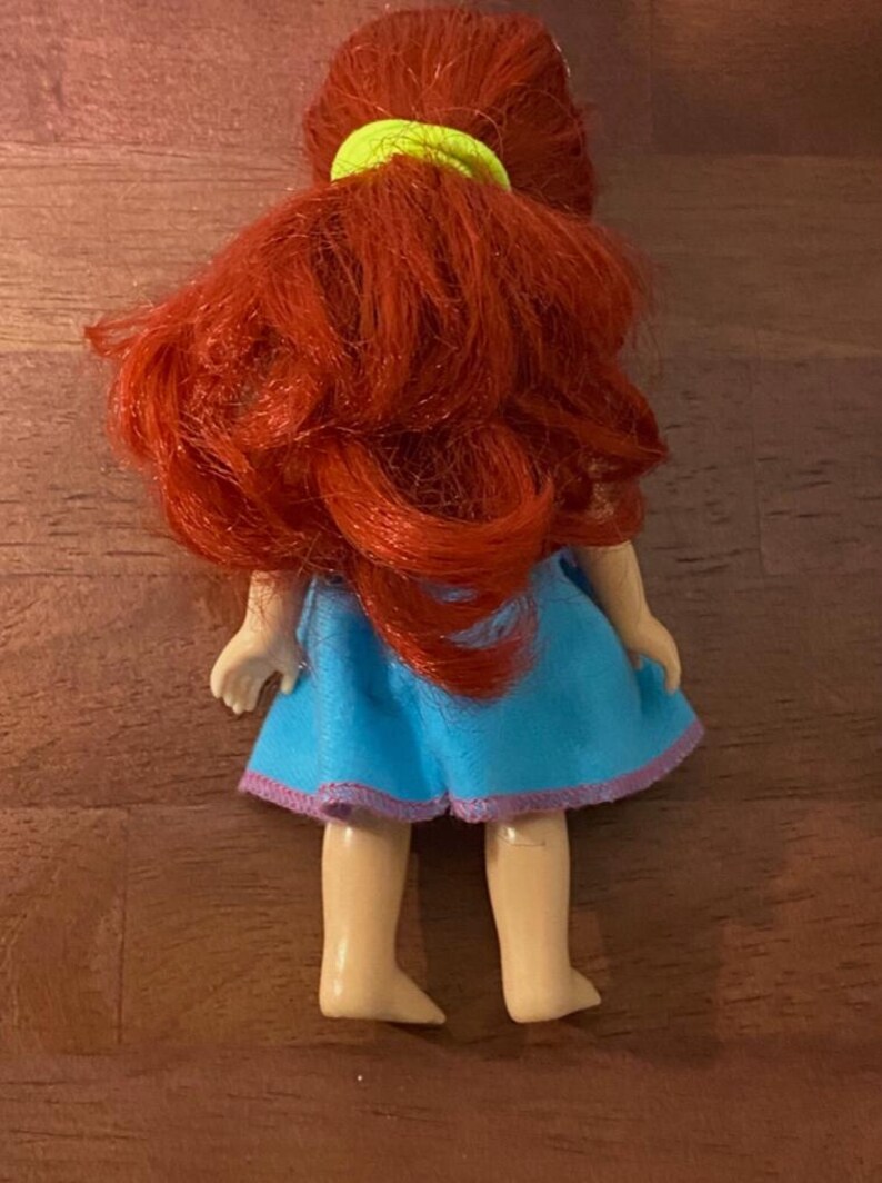 Pode incluir: Uma pequena boneca com cabelo vermelho vivo penteado em cachos soltos, preso com um el&aacute;stico verde n&eacute;on. A boneca usa um vestido azul claro com acabamento roxo. As pernas e os p&eacute;s da boneca s&atilde;o vis&iacute;veis.