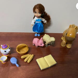 Pode incluir: Uma cole&ccedil;&atilde;o de miniaturas de brinquedos e acess&oacute;rios, incluindo uma boneca com um vestido azul, um porco rosa, uma ovelha amarela, um cavalo, uma chaleira, uma cesta, p&atilde;o, uma escova de cabelo e um livro aberto. Os brinquedos est&atilde;o dispostos numa superf&iacute;cie de madeira.