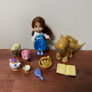 Pode incluir: Uma cole&ccedil;&atilde;o de brinquedos em miniatura com uma boneca em um vestido azul, um cavalo, uma ovelha, um porco, uma chaleira, uma x&iacute;cara, uma escova, uma cesta de p&atilde;o e um livro aberto. Os brinquedos est&atilde;o dispostos em uma superf&iacute;cie de madeira.