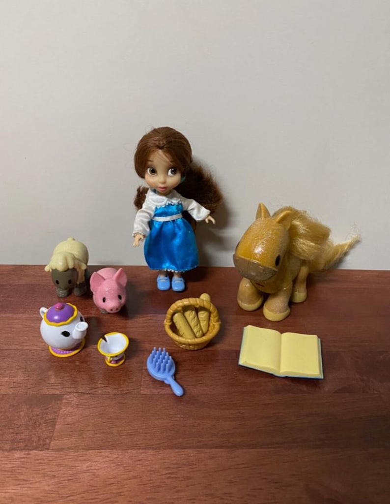 Pode incluir: Uma cole&ccedil;&atilde;o de brinquedos em miniatura com uma boneca em um vestido azul, um cavalo, uma ovelha, um porco, uma chaleira, uma x&iacute;cara, uma escova, uma cesta de p&atilde;o e um livro aberto. Os brinquedos est&atilde;o dispostos em uma superf&iacute;cie de madeira.