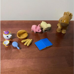 Pode incluir: Uma cole&ccedil;&atilde;o de pequenas figuras de brinquedo e acess&oacute;rios coloridos. Inclui um porco rosa, uma ovelha amarela, um cavalo castanho, uma chaleira branca com tampa roxa, uma cesta amarela, um livro azul, uma escova azul e p&atilde;o amarelo.