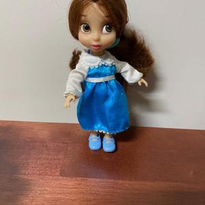 Pode incluir: Uma pequena boneca de uma personagem com cabelo castanho e olhos grandes, vestida com um vestido azul com gola e mangas brancas. A boneca tem sapatos azuis e est&aacute; pousada numa superf&iacute;cie castanha.