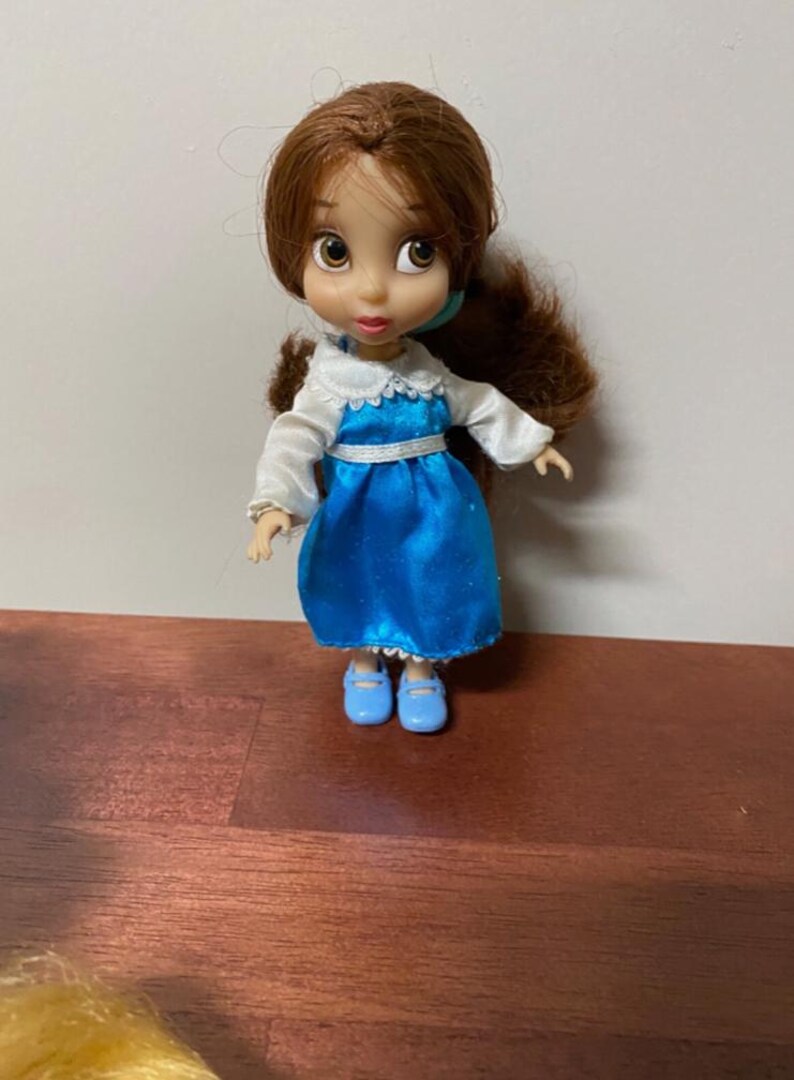 Pode incluir: Uma pequena boneca de uma personagem com cabelo castanho e olhos grandes, vestida com um vestido azul com gola e mangas brancas. A boneca tem sapatos azuis e est&aacute; pousada numa superf&iacute;cie castanha.