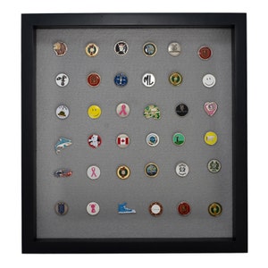 May include: A black framed shadow box with a gray background displaying a collection of 40 golf ball markers. The markers are arranged in a grid pattern and feature a variety of designs, including logos, symbols, and images.