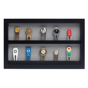 May include: A black wooden display case with a gray felt lining holds ten golf ball markers. The markers are made of metal and feature various designs, including a golf ball, a golfer, and a hole-in-one.