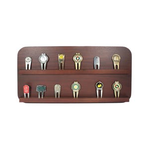 May include: A wooden display case holds twelve golf ball markers. The markers are silver and gold-toned, with various designs and colors. The display case is a dark brown color.