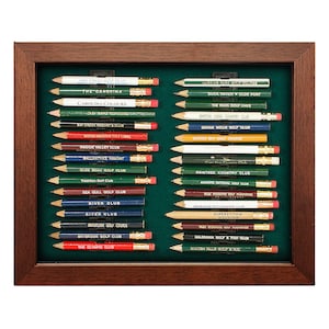 May include: A framed display of 20 golf pencil sets. Each pencil is decorated with the name of a different golf course. The pencils are arranged in two rows, with the names of the courses facing the viewer. The frame is made of dark wood and has a green felt backing.