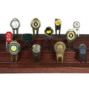 May include: A collection of 13 golf ball markers and divot tools displayed on a tiered wooden stand. The markers feature various designs, including logos and images.