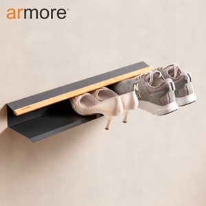 Op de afbeelding: Een grijs metalen schoenenrek met een houten plank en een zwart metalen frame. Het rek is aan een muur gemonteerd en heeft twee paar schoenen erop, een paar witte sneakers en een paar beige hakken.