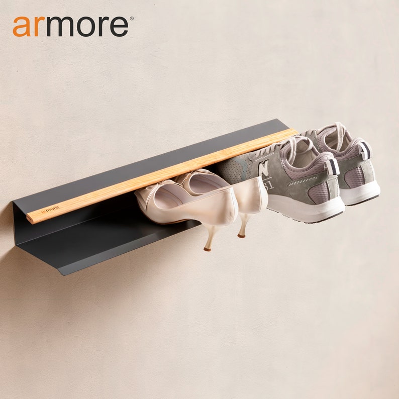 Op de afbeelding: Een grijs metalen schoenenrek met een houten plank en een zwart metalen frame. Het rek is aan een muur gemonteerd en heeft twee paar schoenen erop, een paar witte sneakers en een paar beige hakken.