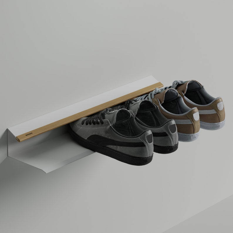 Op de afbeelding: Drie paar sneakers, twee grijs en een bruin, worden getoond op een wit rek met een houten rail. Het rek is gemonteerd aan een witte muur. De tekst "SENSO" is zichtbaar op de rail.