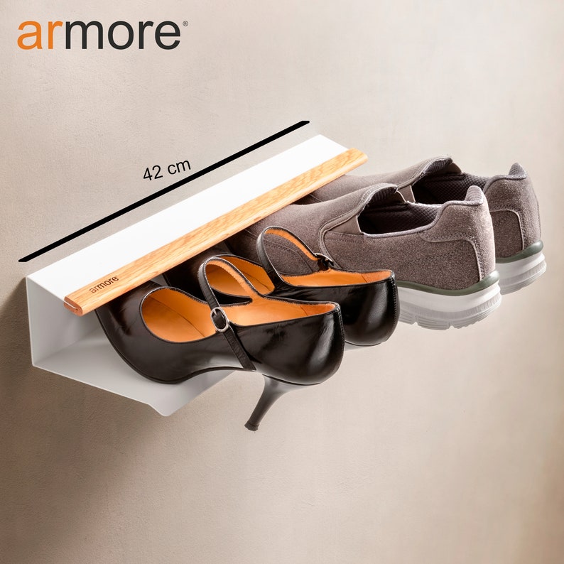 Op de afbeelding: Een wit, aan de muur gemonteerd schoenenrek met een houten plank. Het rek is 42 cm lang en biedt ruimte voor drie paar schoenen, waaronder een paar zwarte hakken en een paar grijze sneakers. Het rek is gelabeld met "armore".