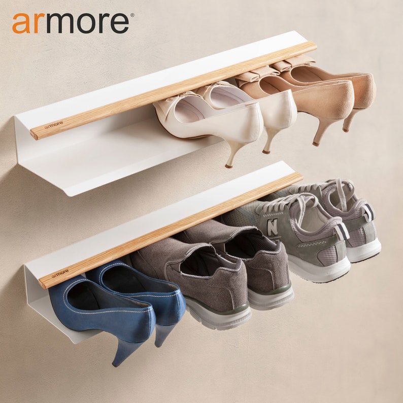 Op de afbeelding: Witte, aan de muur gemonteerde schoenenrek met twee planken en een houten rail. De bovenste plank heeft drie paar witte hakken en de onderste plank heeft twee paar grijze sneakers en een paar blauwe hakken.