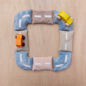 Op de afbeelding: Een houten speelgoedauto in geel en oranje staat op een speelweg van blauwe, beige en roze stoffen stukken. De wegdelen zijn in een vierkante vorm op een licht houten oppervlak gerangschikt.