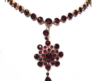 Op de afbeelding: Een gouden ketting met een hanger met een cluster van rode edelstenen. De hanger heeft de vorm van een bloem met een traanvormige edelsteen die aan het midden hangt.