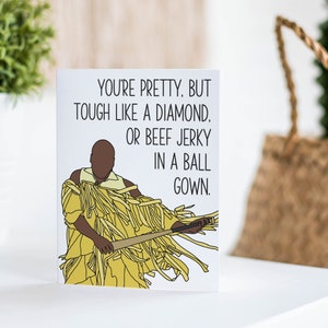 May include: A greeting card with a cartoon illustration of a person wearing a yellow and brown outfit. The text on the card reads "You're pretty. But tough like a diamond. Or beef jerky in a ball gown."