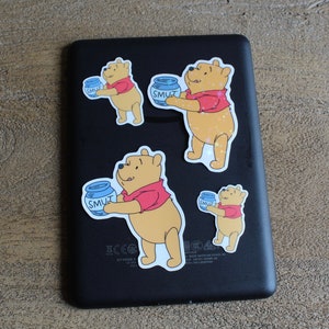 May include: Four Winnie the Pooh stickers on a black background. Each sticker features Winnie the Pooh holding a blue jar with the word "SMUT" on it. The stickers are different sizes and are arranged in a random pattern.