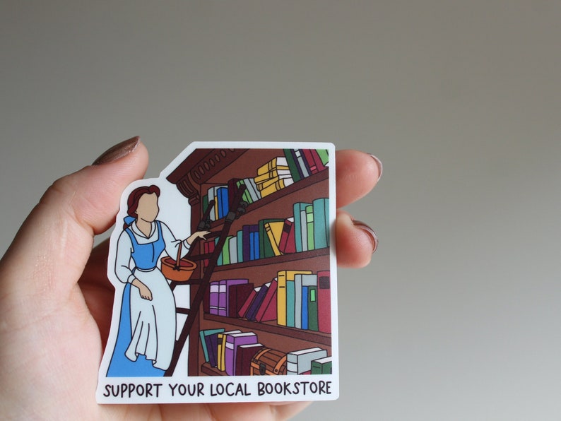 May include: A sticker featuring a cartoon illustration of Belle from Beauty and the Beast reaching for a book on a bookshelf. The sticker has the text "Support Your Local Bookstore" at the bottom.