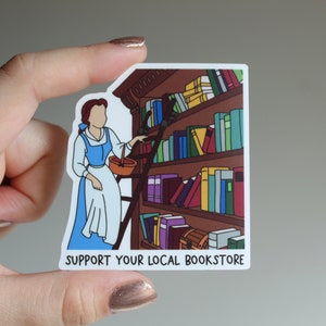 May include: A sticker featuring a cartoon illustration of Belle from Beauty and the Beast reaching for a book on a bookshelf. The sticker has the text "SUPPORT YOUR LOCAL BOOKSTORE" at the bottom.