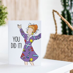 May include: A greeting card with a cartoon illustration of a woman wearing a purple dress with science-themed patterns. The text "You Did It!" is printed in black on the card.