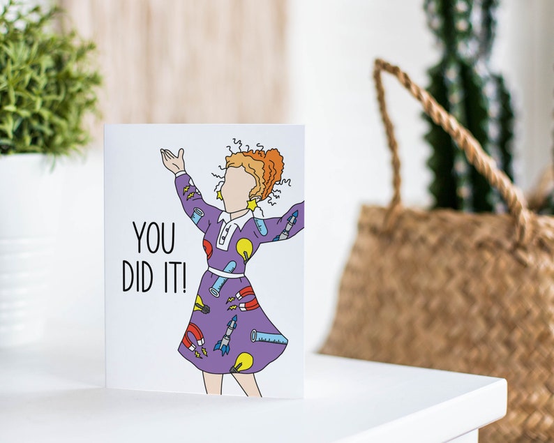 May include: A greeting card with a cartoon illustration of a woman wearing a purple dress with science-themed patterns. The text "You Did It!" is printed in black on the card.