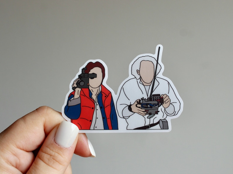 May include: A sticker featuring a line drawing of two characters from the film Back to the Future. The character on the left is holding a video camera and the character on the right is holding a device with wires.
