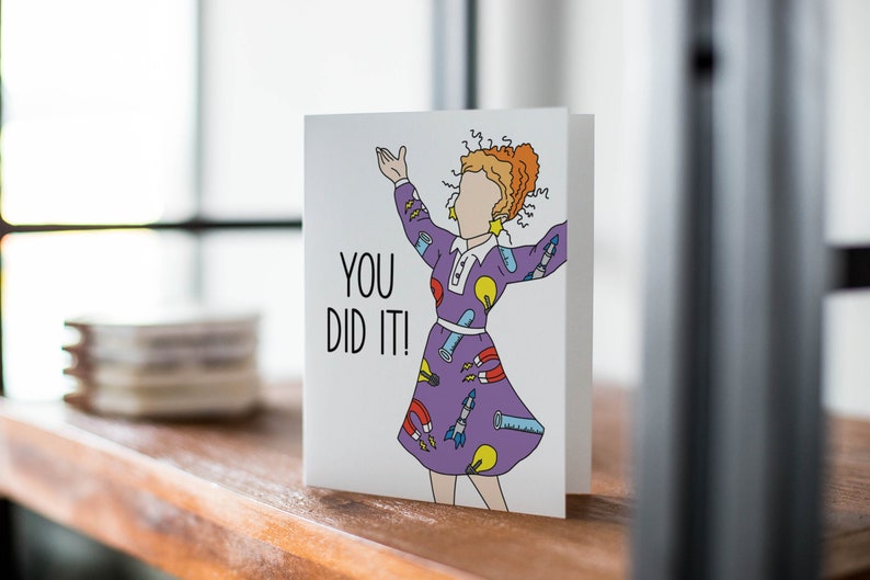 May include: A white card with a cartoon illustration of a person wearing a purple dress with science-themed patterns. The text "YOU DID IT!" is printed on the card.