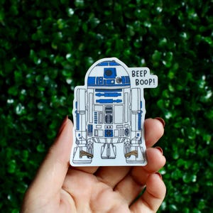 Può includere: Un adesivo raffigurante il droide R2-D2 di Star Wars in blu, bianco e argento. L'adesivo ha il testo "BEEP BOOP!" in un fumetto. Lo sfondo è una siepe verde.